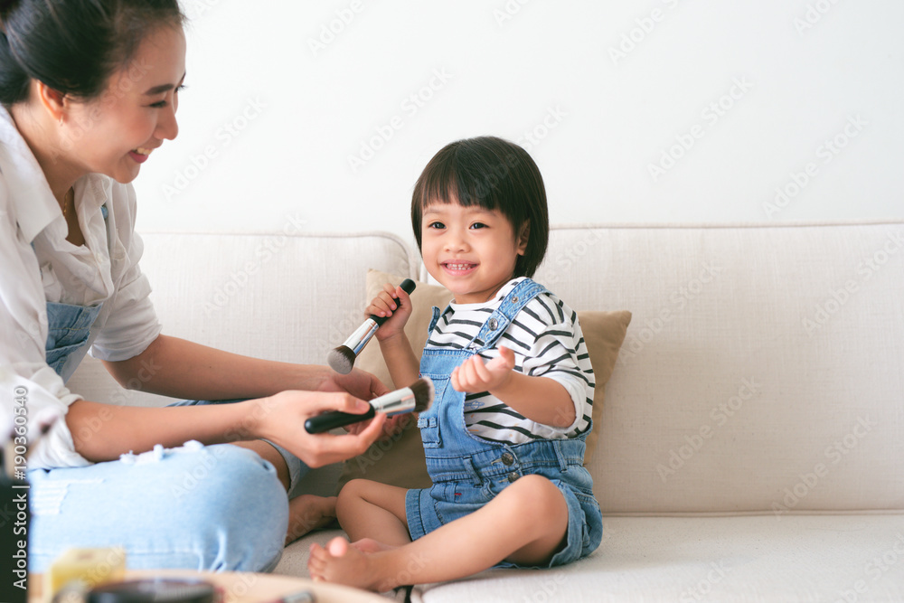 可爱的小女孩和她漂亮的妈妈坐在家里的沙发上化妆