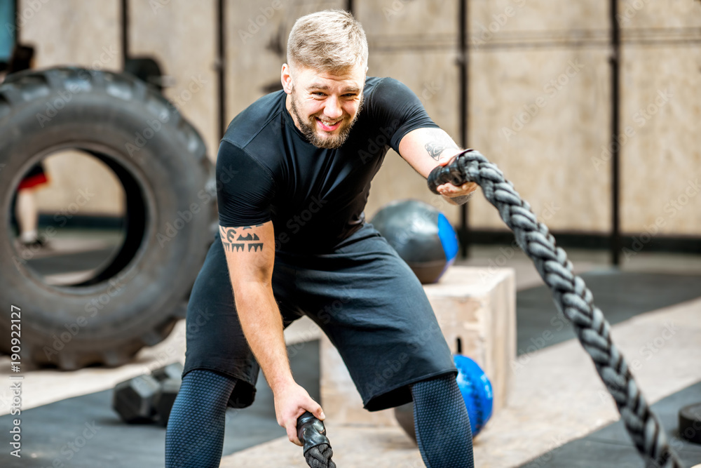 Athletic man in black sportswear swinging with a big rope training in the gym