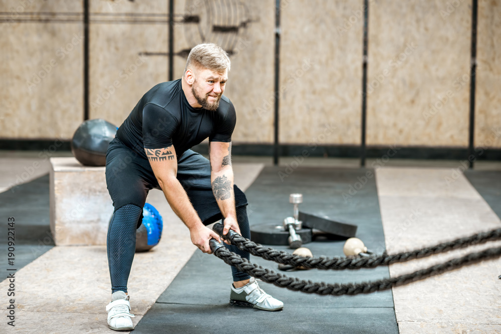 Athletic man in black sportswear swinging with a big rope training in the gym