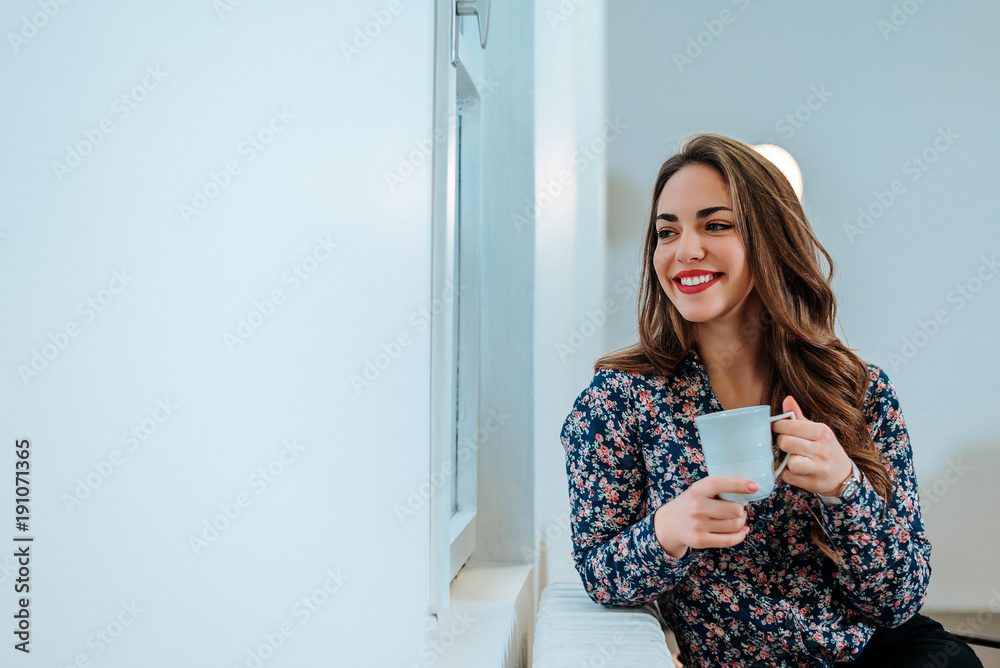 Portrait of beautiful woman looking through the window holding cup of tea.