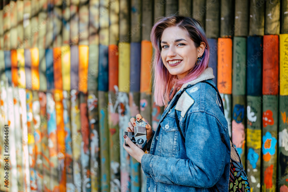 Smiling  young woman with dyed hair holding camera near the hand painted wall.Smiling young woman wi