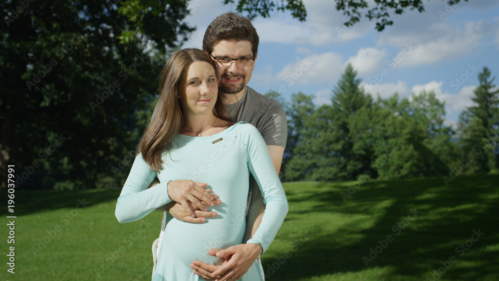 PORTRAIT: Embraced pregnant couple standing in a green meadow on a sunny day