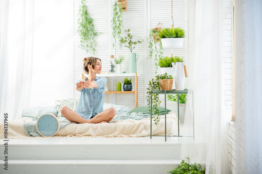 Young woman having a good morning sitting on the bed in the beautiful bright bedroom with green plan