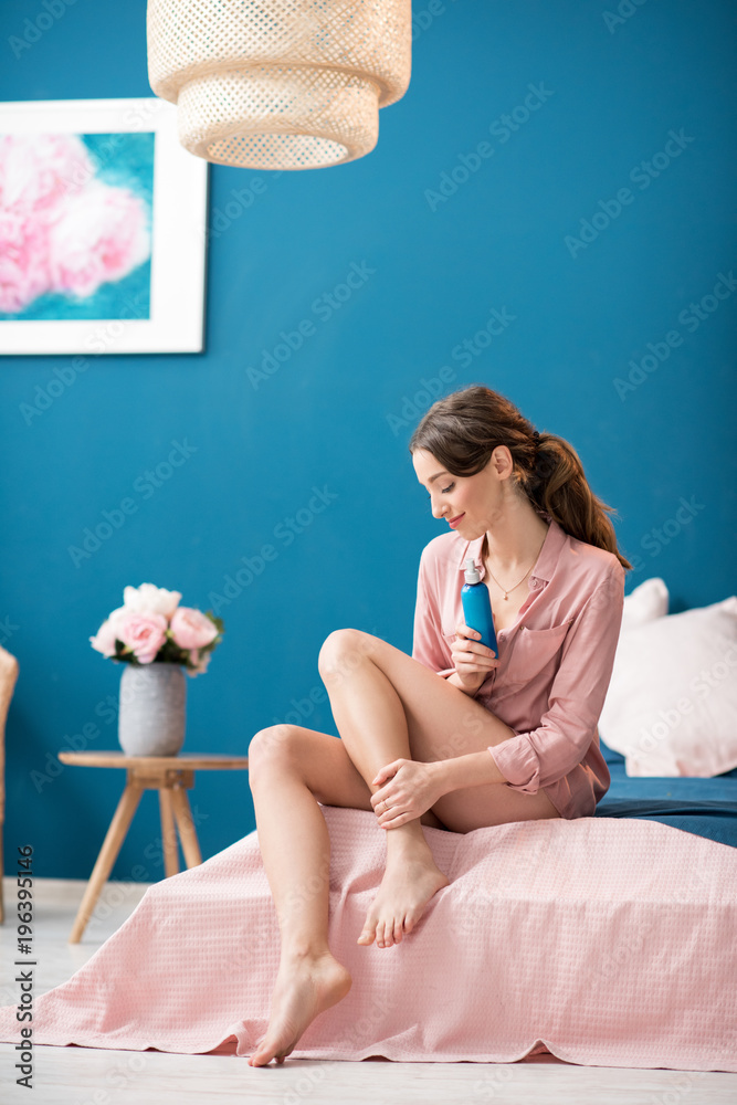 Young woman applying a lotion on her legs sitting on the bed in the beautiful pink and blue bedroom 