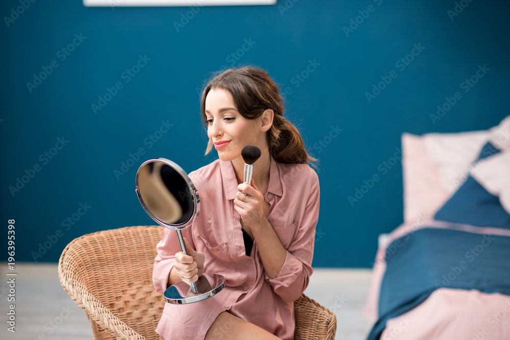 Young woman in pink shirt applying cosmetics with brush sitting on the chair in the beautiful blue b