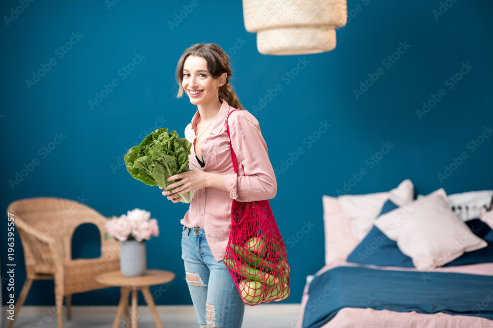 Portrait of a young woman standing with red bag full of green vegetables holding a lettuce in the bl