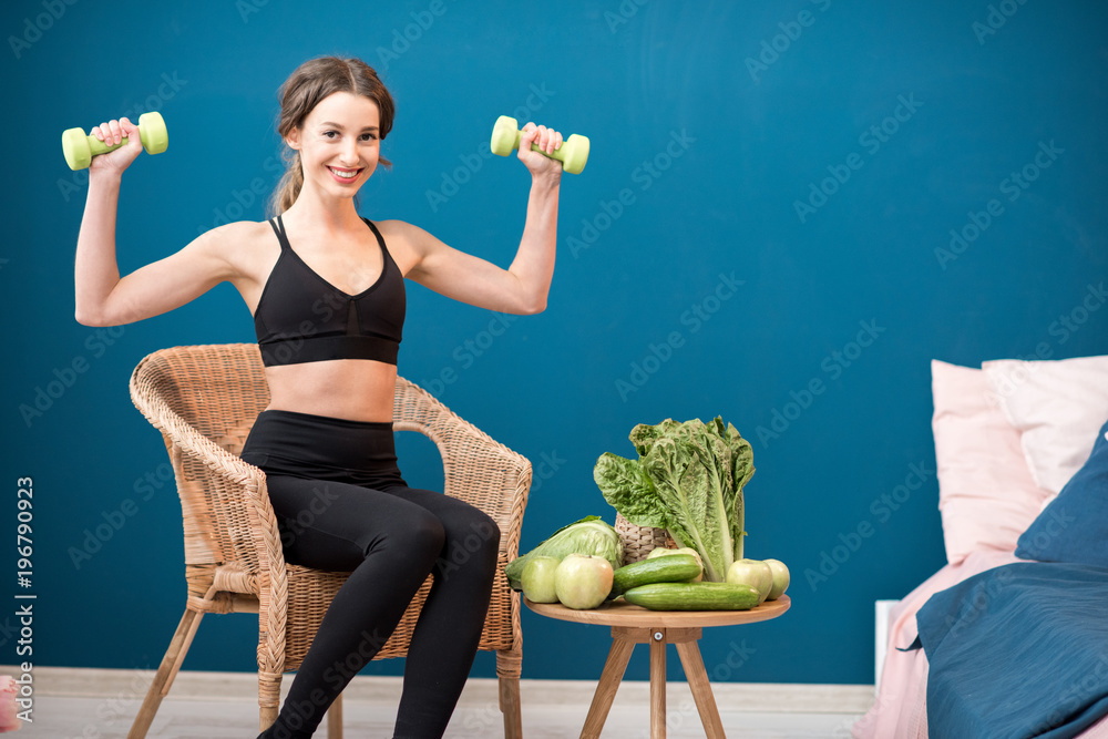 Portrait of a young sports woman sitting with dumbbells and healthy green food indoors on the blue w