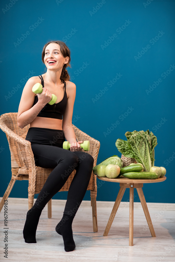 Portrait of a young sports woman sitting with dumbbells and healthy green food indoors on the blue w