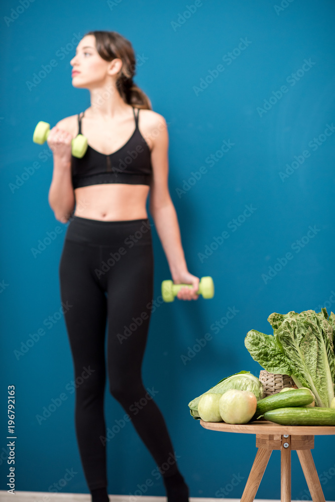 Young woman exercising indoors with green healthy food on the table on the blue wall background at h