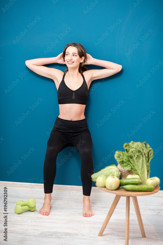 Young woman exercising indoors with green healthy food on the table on the blue wall background at h