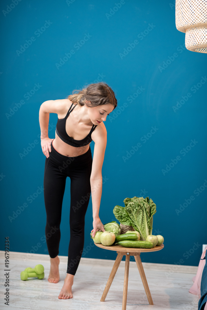 Young woman exercising indoors with green healthy food on the table on the blue wall background at h