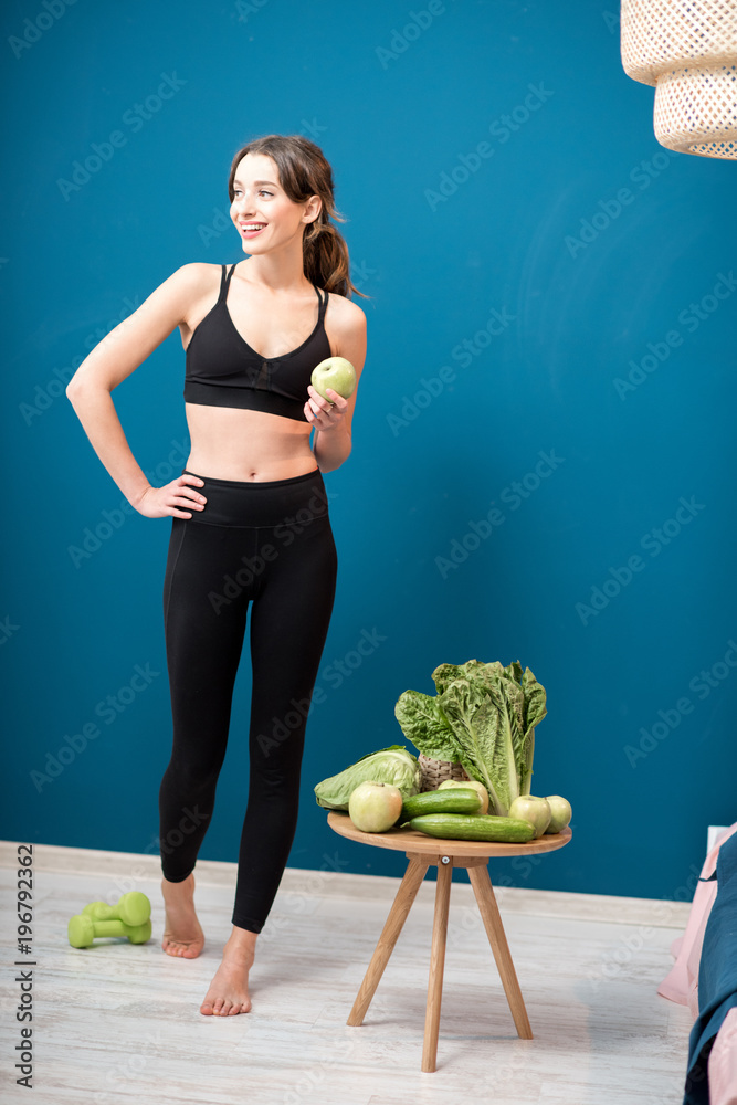 Young woman exercising indoors with green healthy food on the table on the blue wall background at h