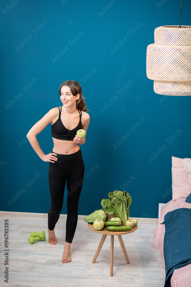 Young woman exercising indoors with green healthy food on the table on the blue wall background at h