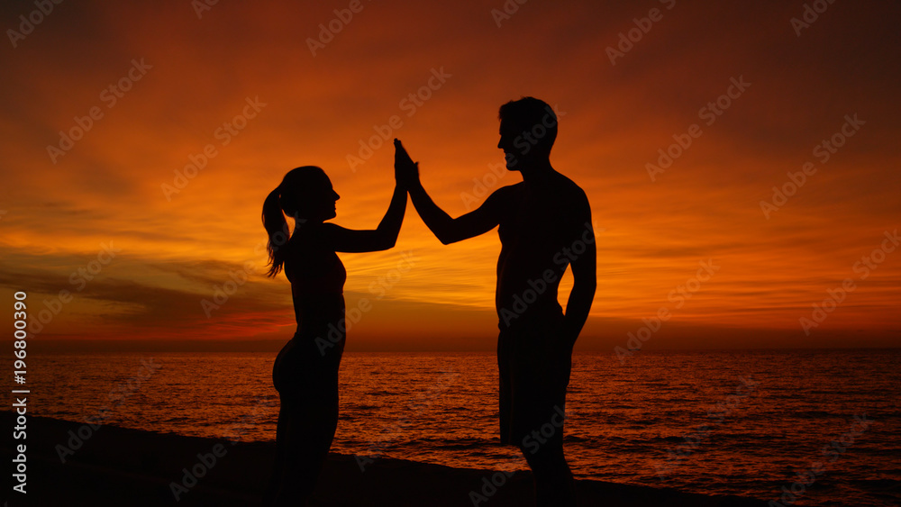 SILHOUETTE: Young man and woman high five by the ocean at sunset after workout.