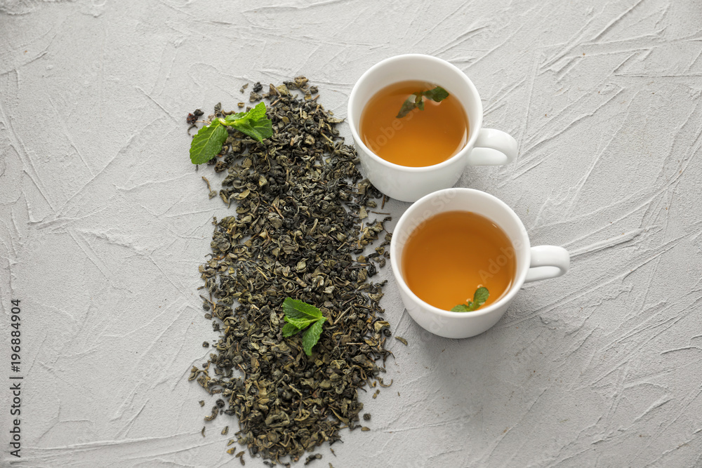 Dried green tea leaves and cups of aromatic drink on light background