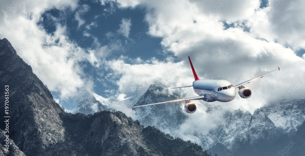 Airplane is flying over low clouds against mountains with snowy peaks in bright day. Landscape. Pass