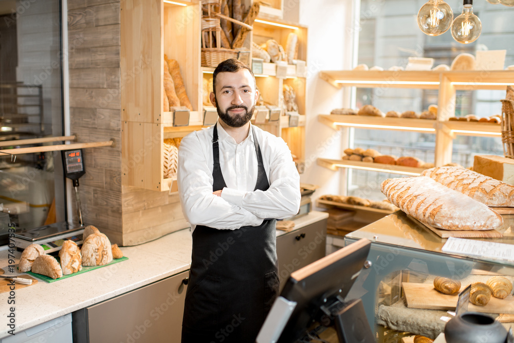 Portrait of a handsome seller in uniform standing in the small and beautiful store with bakery produ