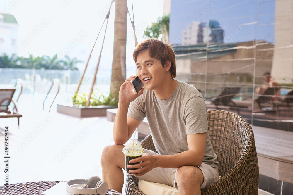 Asian man talking on a phone while sitting outdoors in sunny summer day.