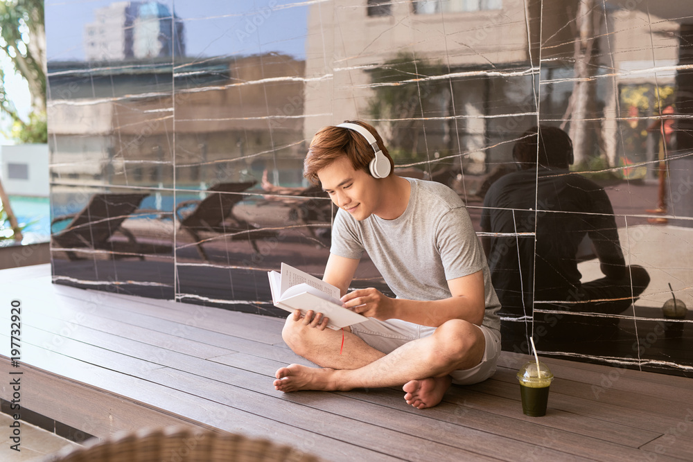 Young asian man reading book and listening to music by the pool on a sunny summer day