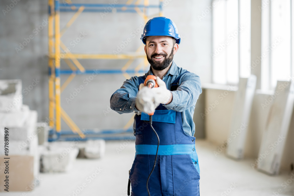 一名身穿工作服的建筑工人在室内建筑工地拿着钻机站着的画像