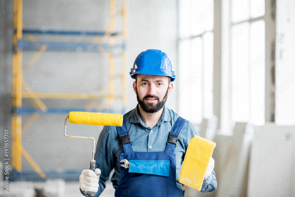 一个穿着工作服的建筑工人在室内拿着黄色油漆卷和研磨工具的肖像