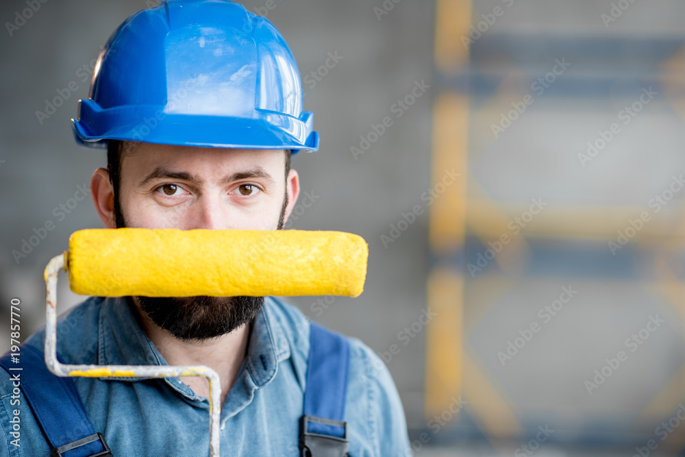 Portrait of a builder in working uniform holding yellow paint roll and grinding tool indoors