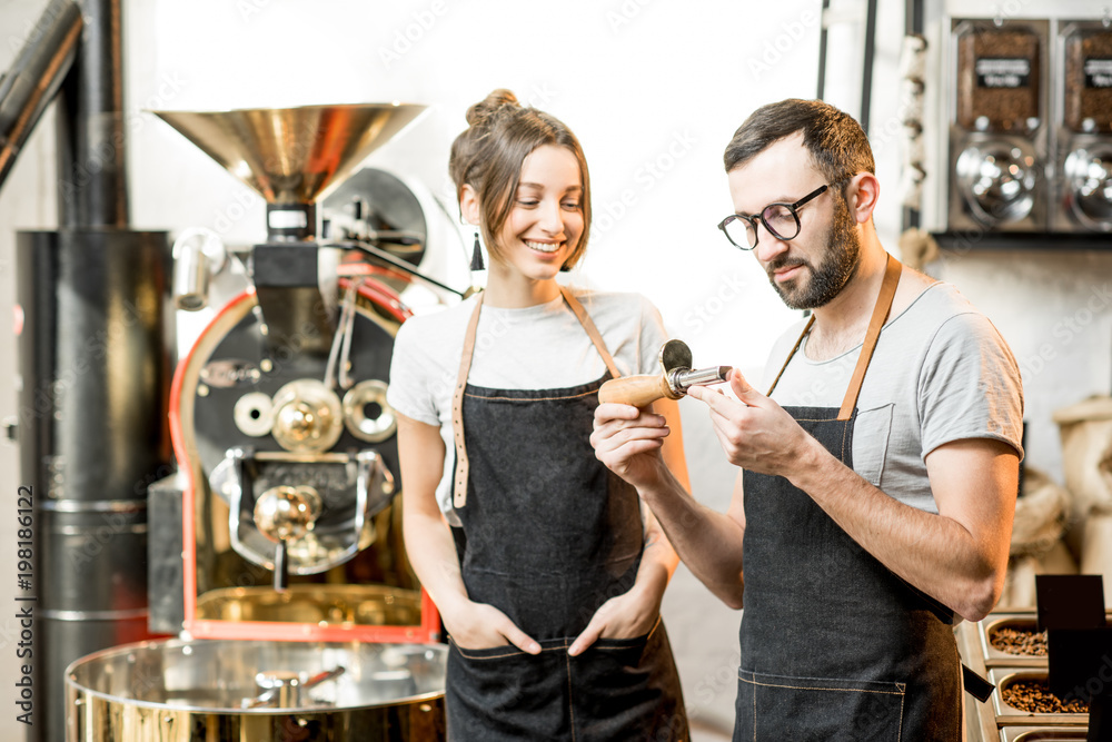 Couple of baristas in uniform checking the quality of roasted coffee beans standing near the roaster