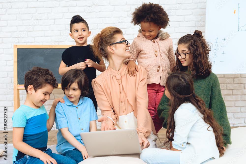 Nice teacher sits with school children who have surrounded her and are looking at laptop.