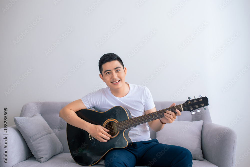 Handsome asian man playing on the guitar on the couch at home