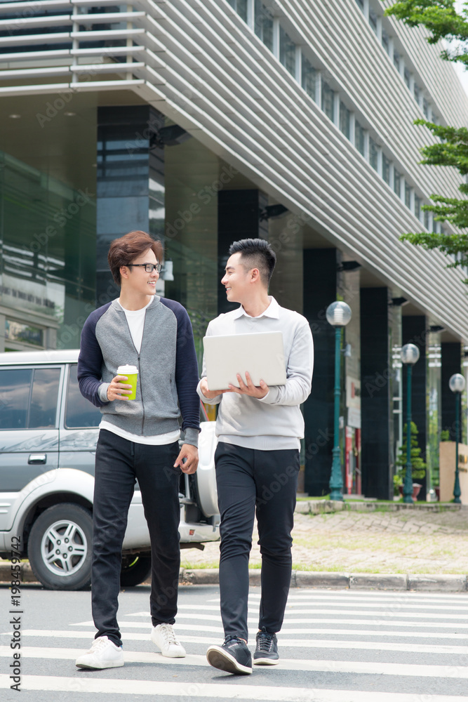 Two smiling young businessmen walking and talking in the city