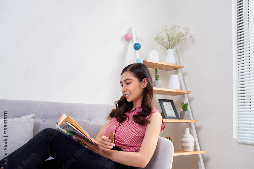 Young beautiful asian woman sitting on couch reading a book enjoying her tea in living room at home