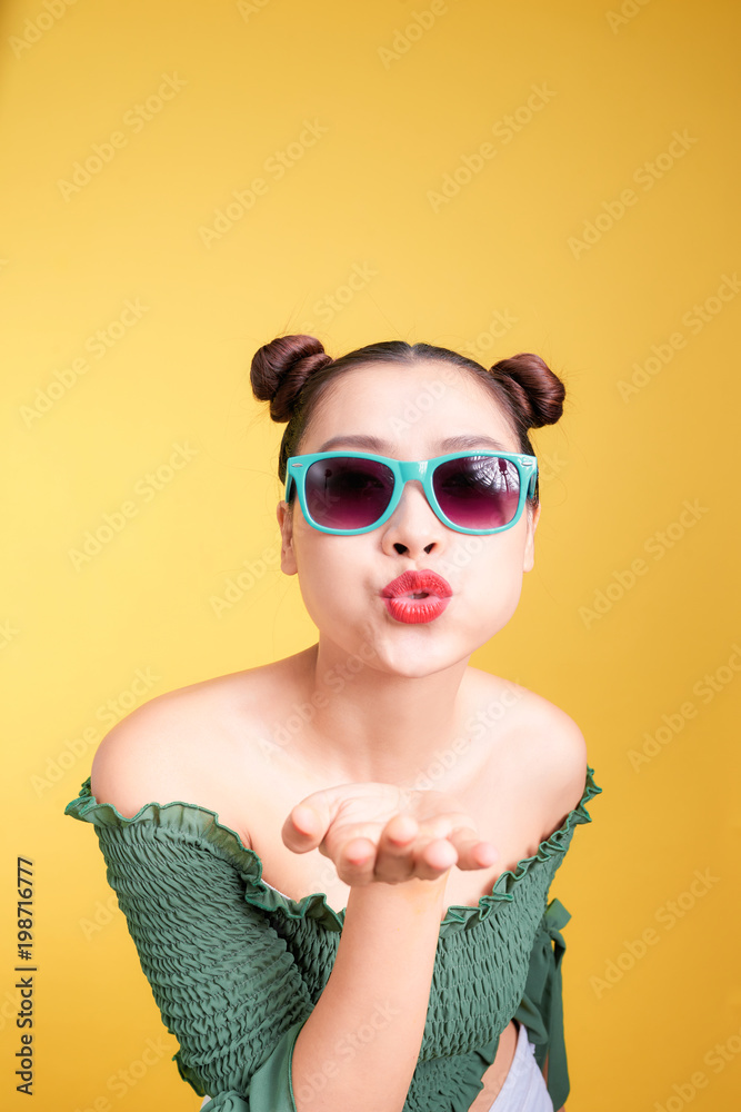 Fashionable asian woman in trendy sunglasses sends a kiss against bright yellow background