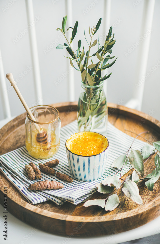 Healthy vegan turmeric latte or golden milk with honey in striped cup on wooden tray, selective focu