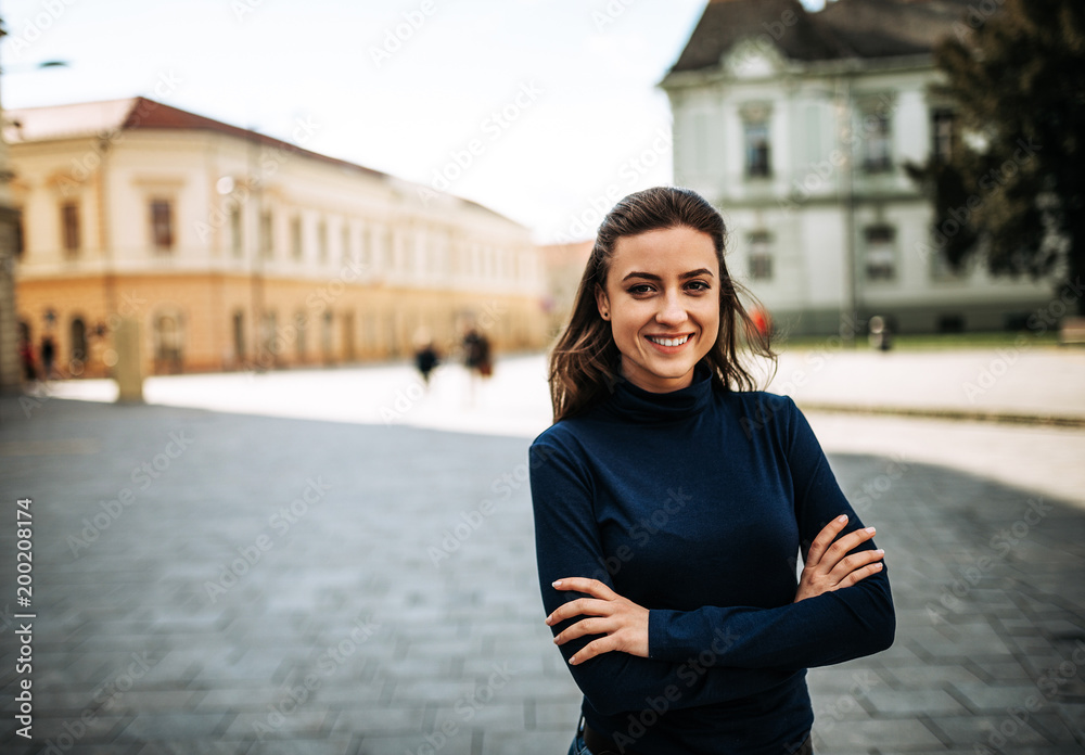 一位双臂交叉、面带微笑的年轻女子站在城市中的肖像。