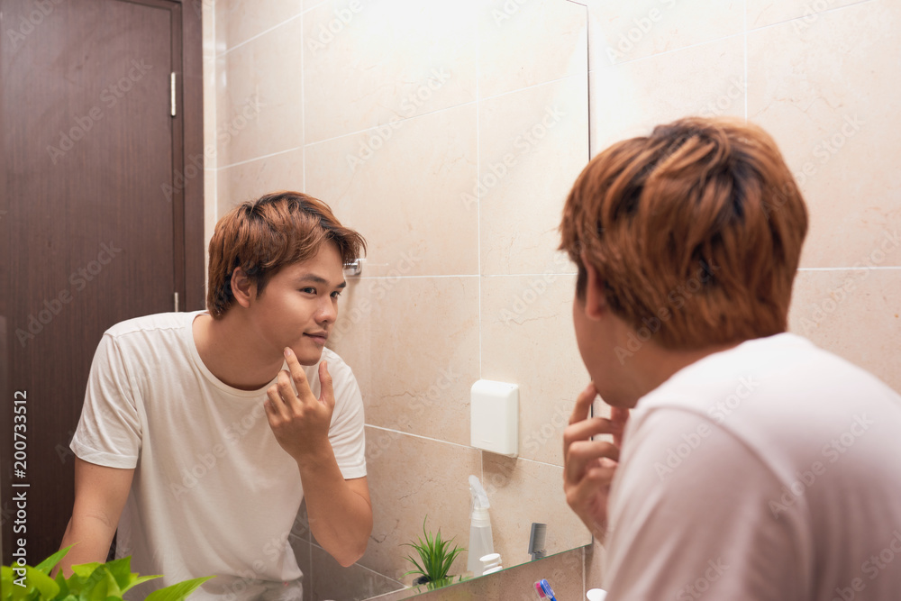 Young asian man looking in mirror and checking his skin in bathroom