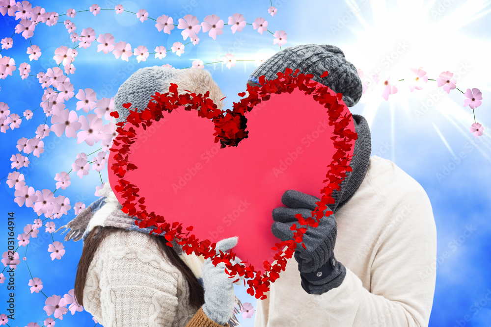 Attractive young couple in warm clothes holding red heart against digitally generated pretty flower 