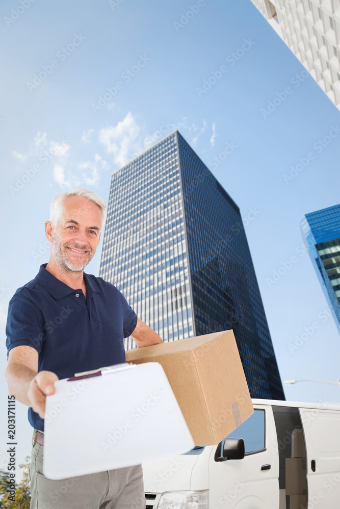 Happy delivery man holding cardboard box and clipboard against skyscraper