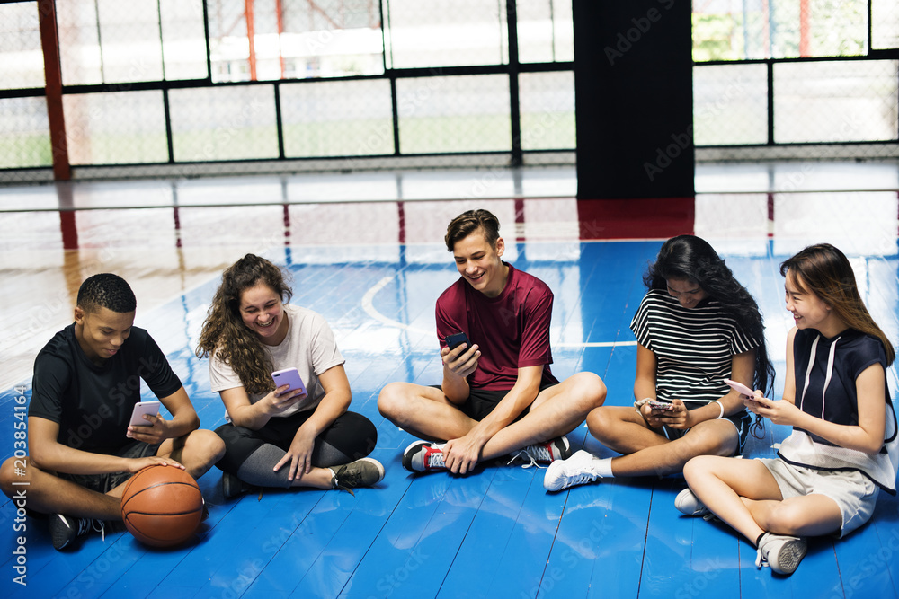 Group of young teenager friends on a basketball court relaxing and using smartphone