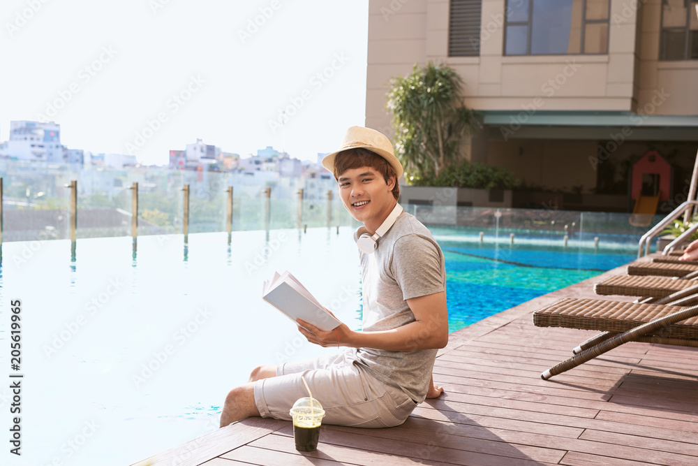 阳光明媚的夏日，一名亚洲年轻男子在泳池边看书