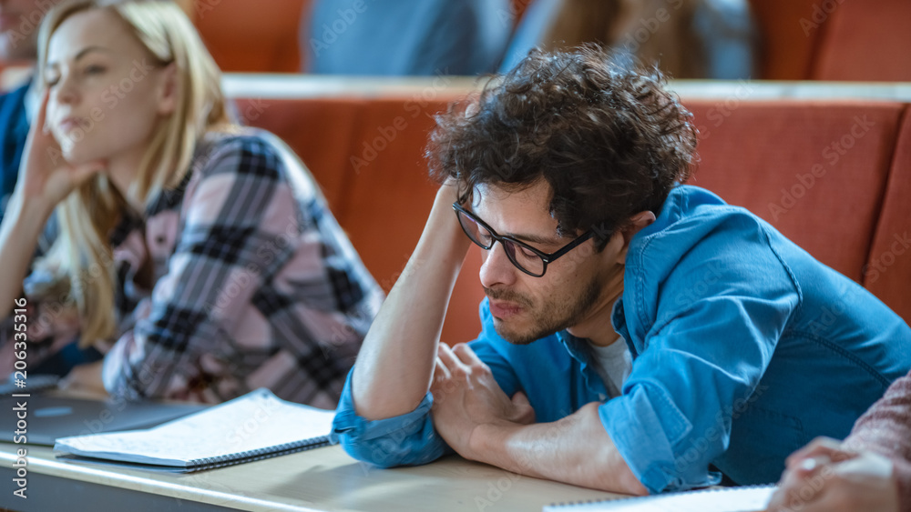 无聊的男学生在大学听讲座。疲惫、疲惫、过度劳累的年轻男霍尔