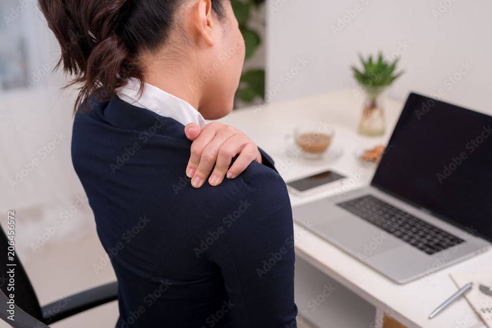 Young business person with neck pain. Focus on hand on neck with laptop on table in background.