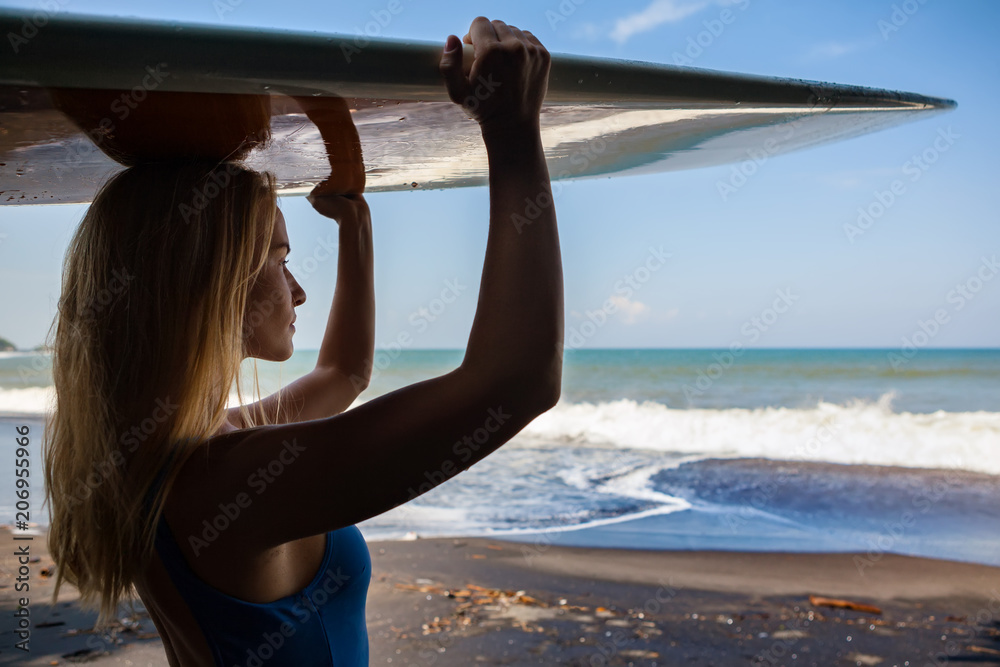 Girl in bikini with surfboard walk on black sand beach. Surfer woman look at sea surf and water pool