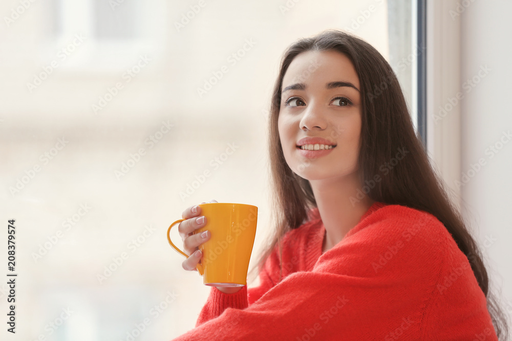 漂亮的年轻女人在家里靠窗的地方喝着一杯热茶