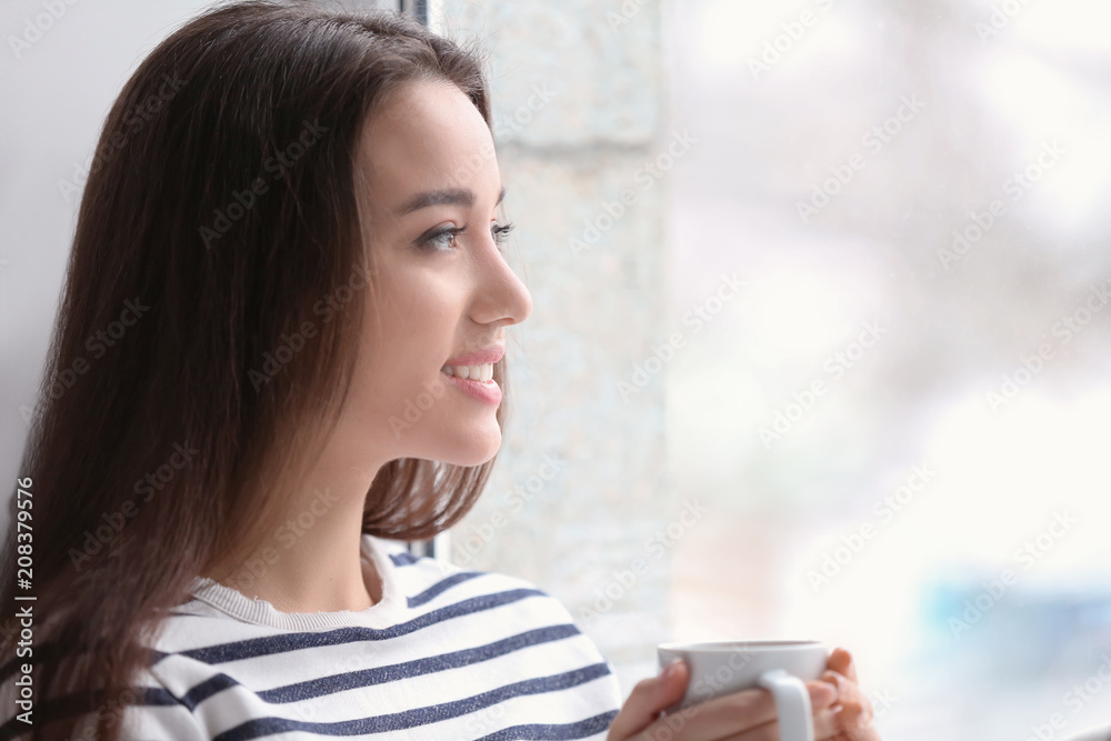 漂亮的年轻女人在家里靠窗喝着热茶