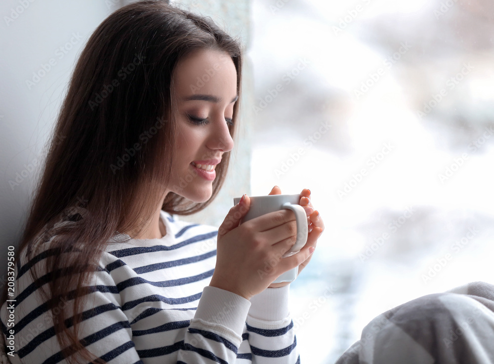 美丽的年轻女子在家里靠窗的地方喝着一杯热茶