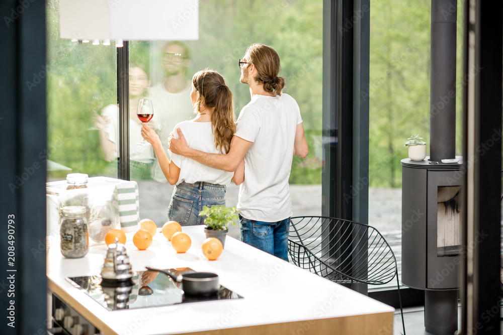 Young couple standing together with wine near the window of the modern apartment enjoying green area