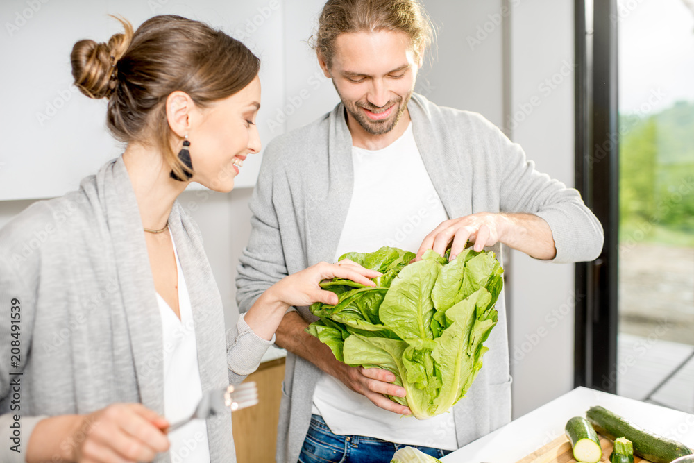 Young cheerful couple dressed alike in gray sweaters cooking some veggie food at the modern kitchen