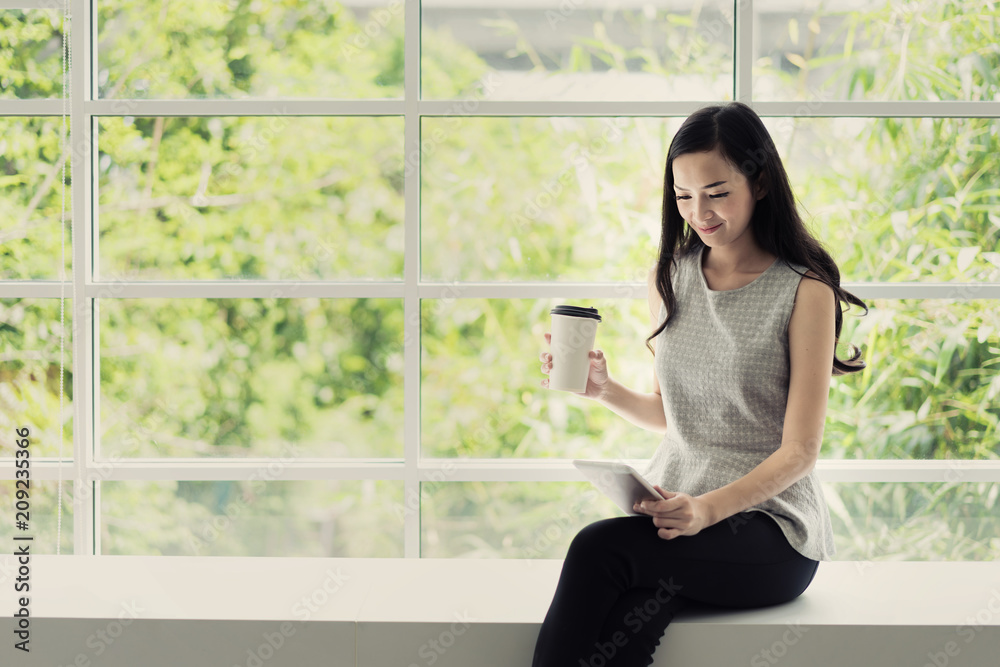 年轻的亚洲微笑女子在咖啡馆喝咖啡，手里拿着一台平板电脑。