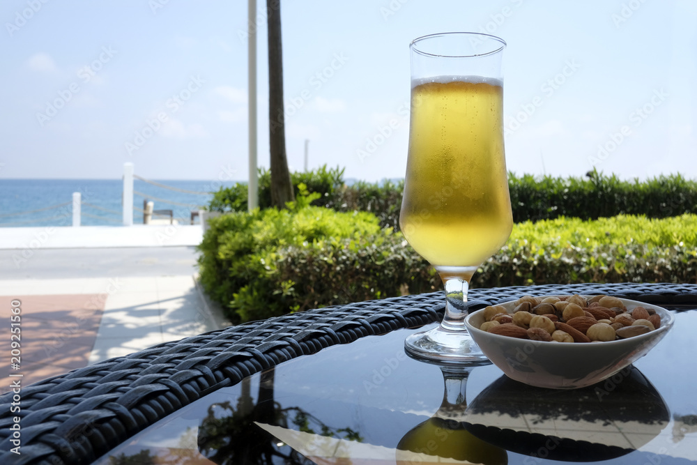 Cup of beer with nuts snacks on glass table. Sunny day near mediterranean sea. Summer time