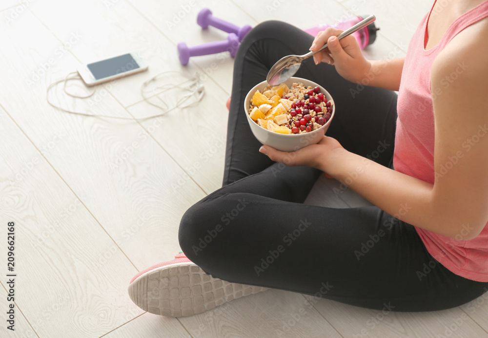 Sporty young woman sitting on floor with bowl of tasty oatmeal